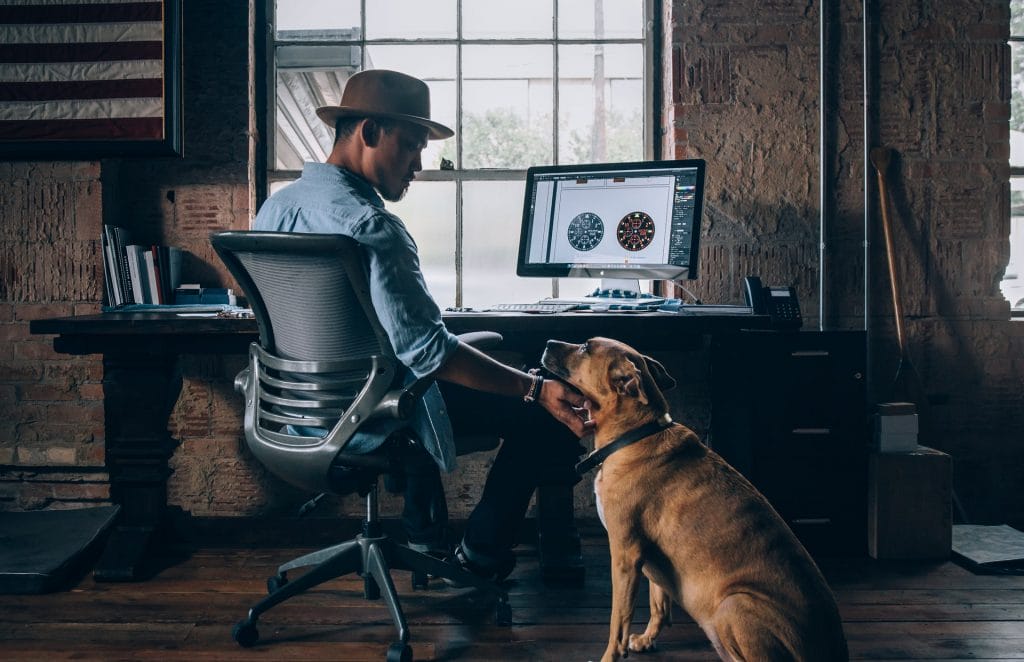 A man sitting at his desk with a hat on, interacting with his dog while looking at his computer screen in a casual office environment.