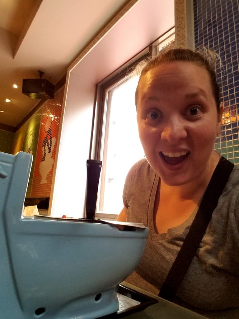 A woman, Kimberly Worsham, smiles cheerfully in front of a toilet, featured in the FLUSH coworking member spotlight.