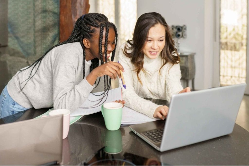 Two young women focus on a laptop, representing the modern work-from-home experience and collaborative efforts.