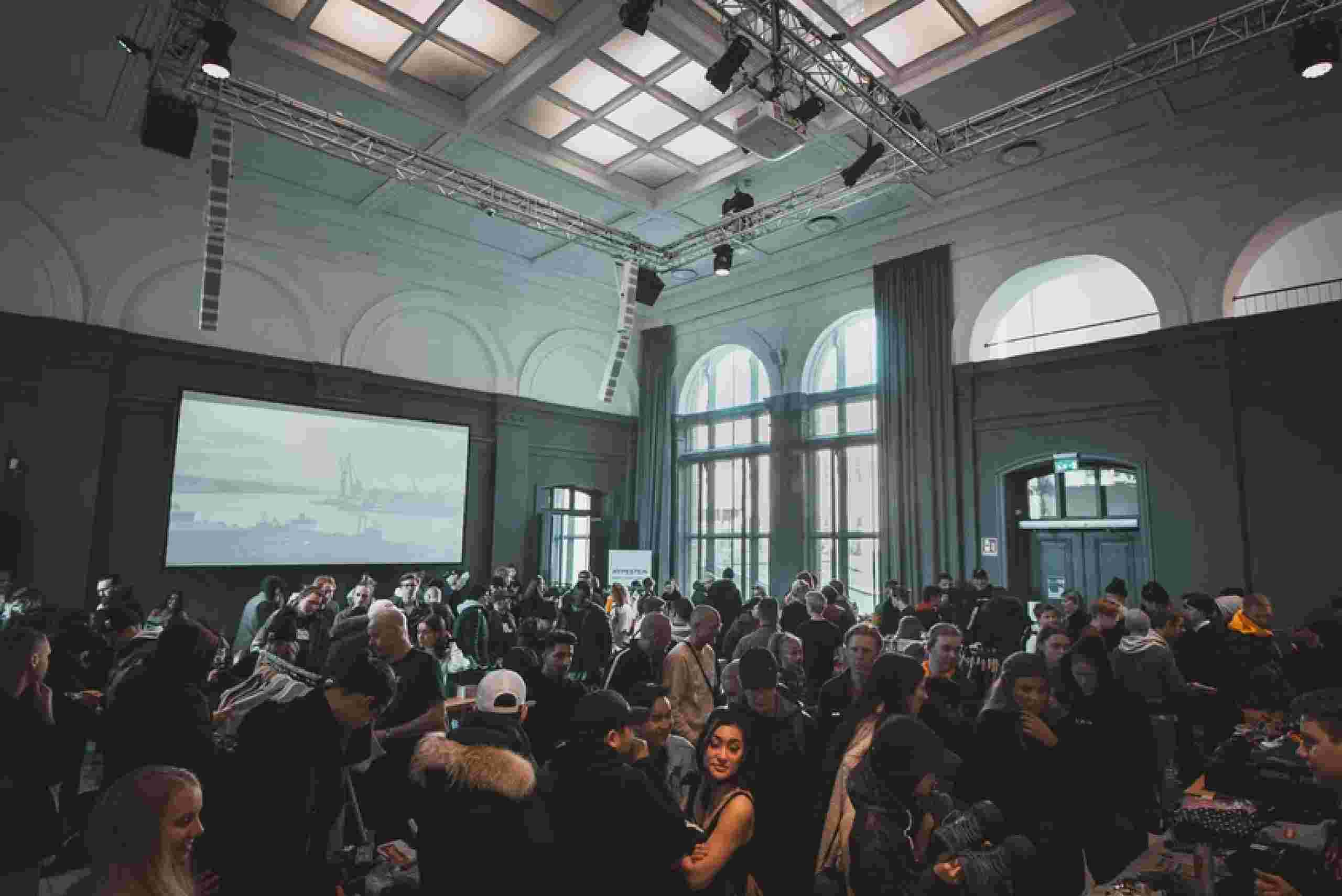 A bustling crowd in a bright room with large windows, actively involved in a corporate gathering.