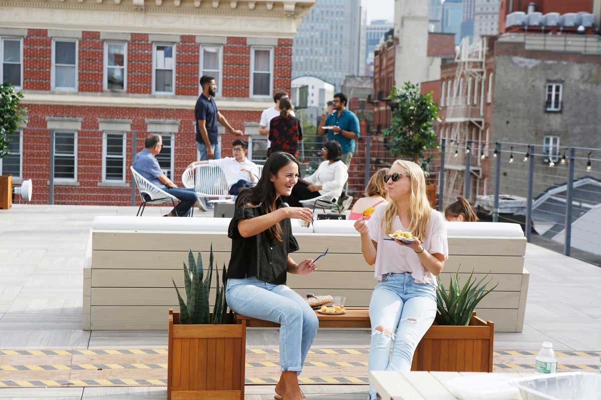 Two women relax on a rooftop, gazing at the cityscape, highlighting a blend of friendship and urban scenery.