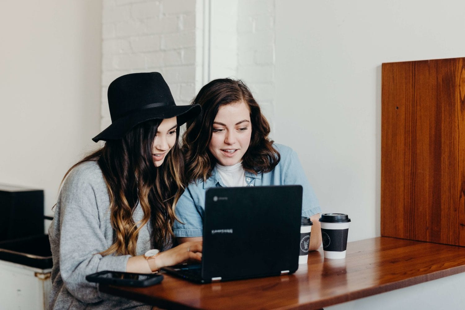  Two women engaged in discussion while seated at a table, focused on a laptop in a coworking environment.