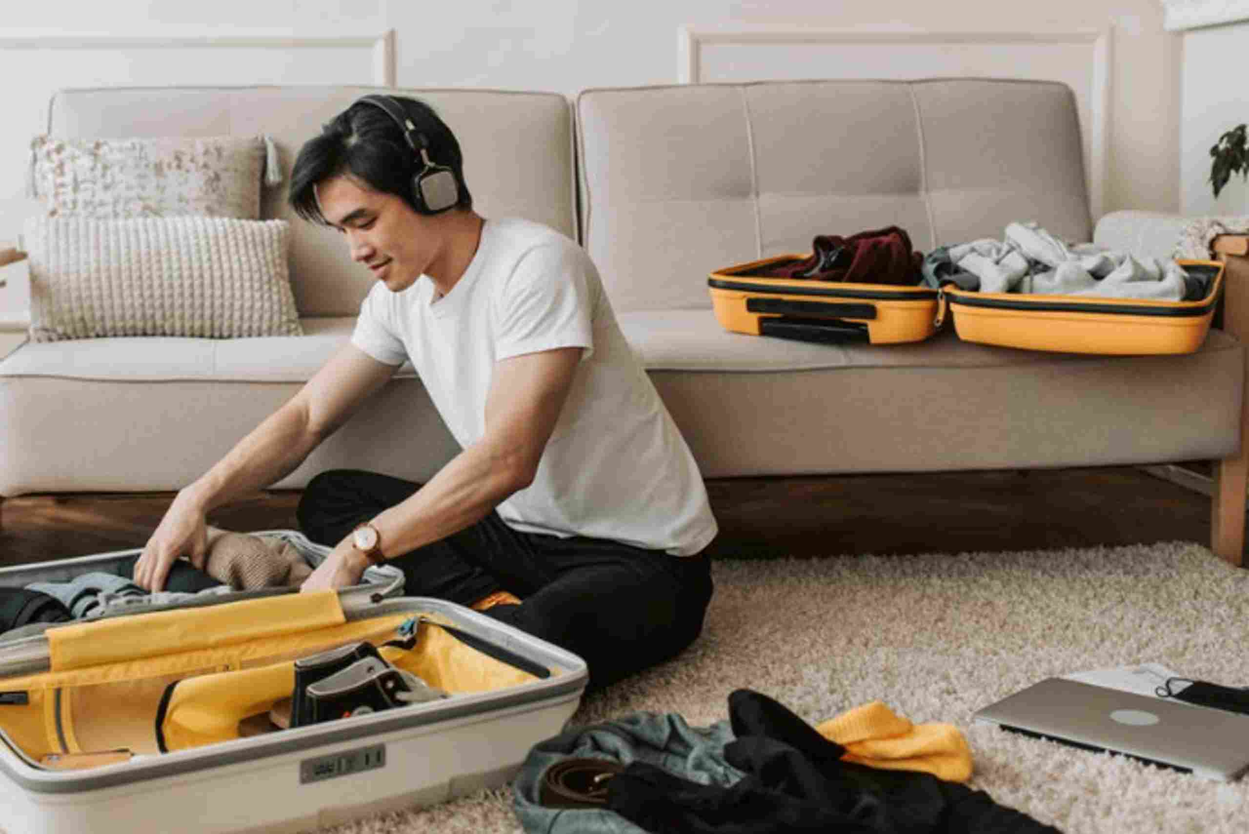 A man sits on the floor with a suitcase, surrounded by essential items for digital nomads, preparing for travel.
