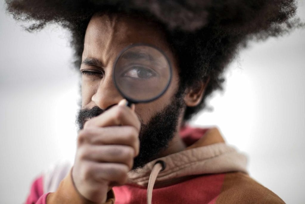 A man with an afro hairstyle inspects an object with a magnifying glass, illustrating the concept of market research.