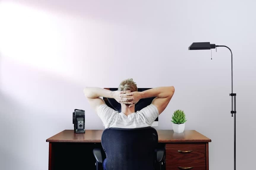 A remote worker relaxes at his desk, arms behind his head, highlighting the comfort of working from home.