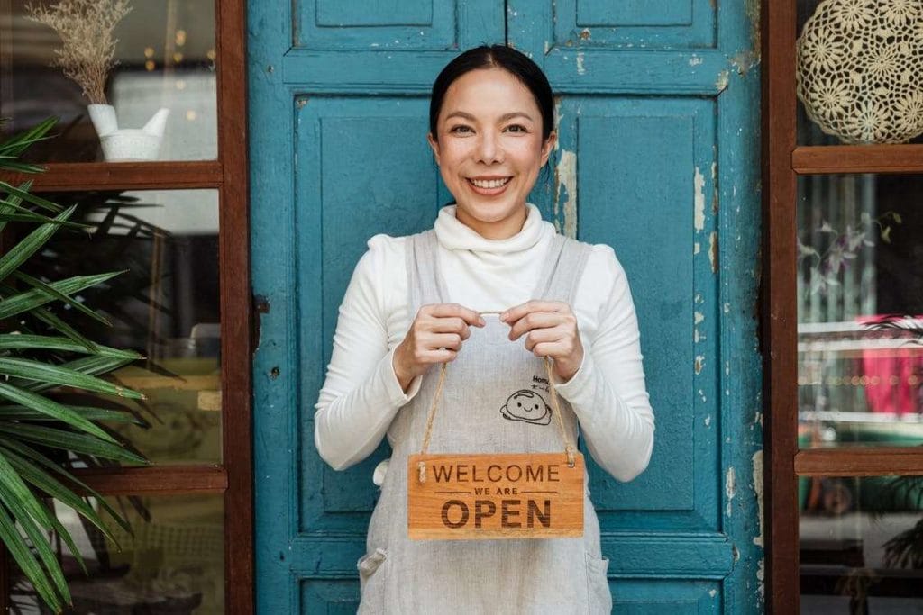 A woman with a welcome sign stands by a door, signaling the start of her online event for small businesses.