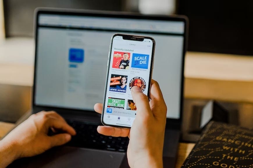 A person browsing podcast options on a smartphone while working on a laptop in a coworking space.