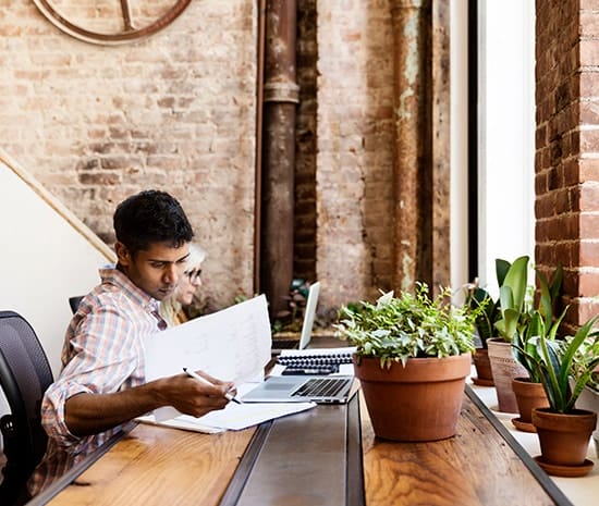 A man at a table with a laptop and papers, focused on work in a coworking environment.